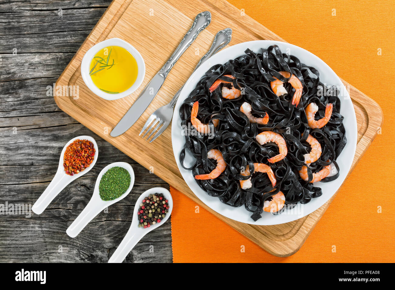 delicious squid ink black noodles with king prawns on white dish on chopping board with olive oil and spices in spoons on background, view from above Stock Photo