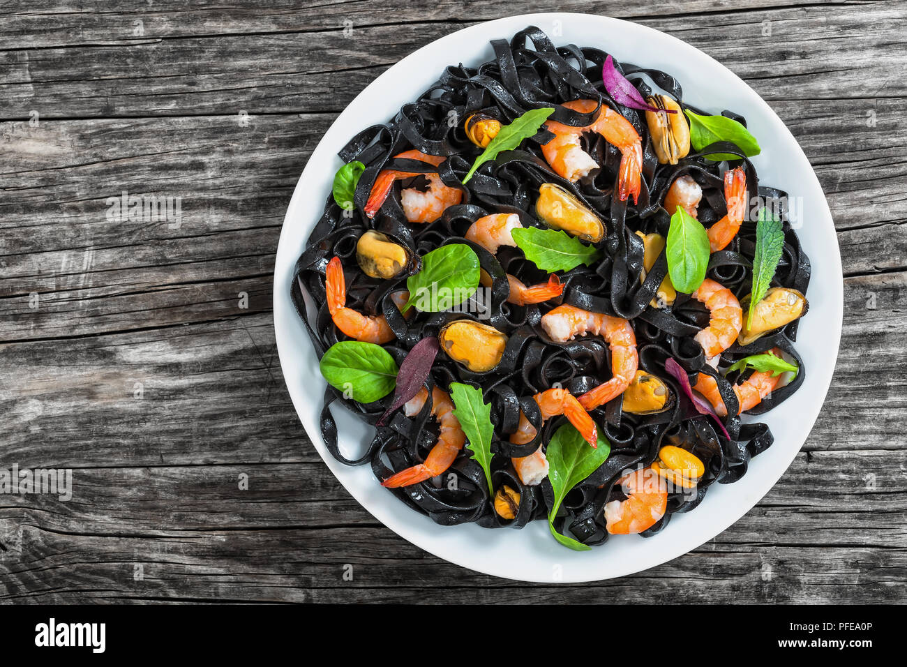 seafood black noodles salad with prawns, mussels, fresh green spinach, lettuce, arugula and mint on white dish on dark wooden table, view from above Stock Photo
