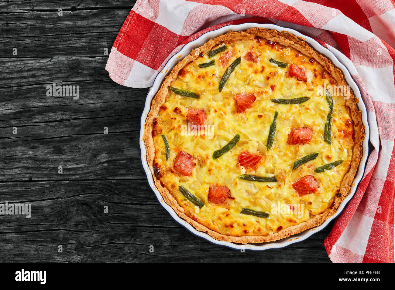 delicious quiche tart with Salmon,green bean and Emmental cheese, cream, onion and eggs in baking dish with kitchen towel on old dark wooden planks, v Stock Photo