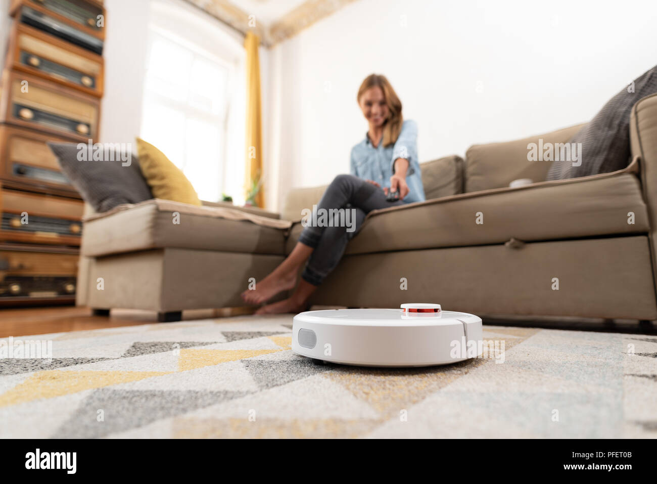 Robotic vacuum cleaner cleaning the room while woman sitting on sofa. Woman controlling vacuum with remote control. Stock Photo