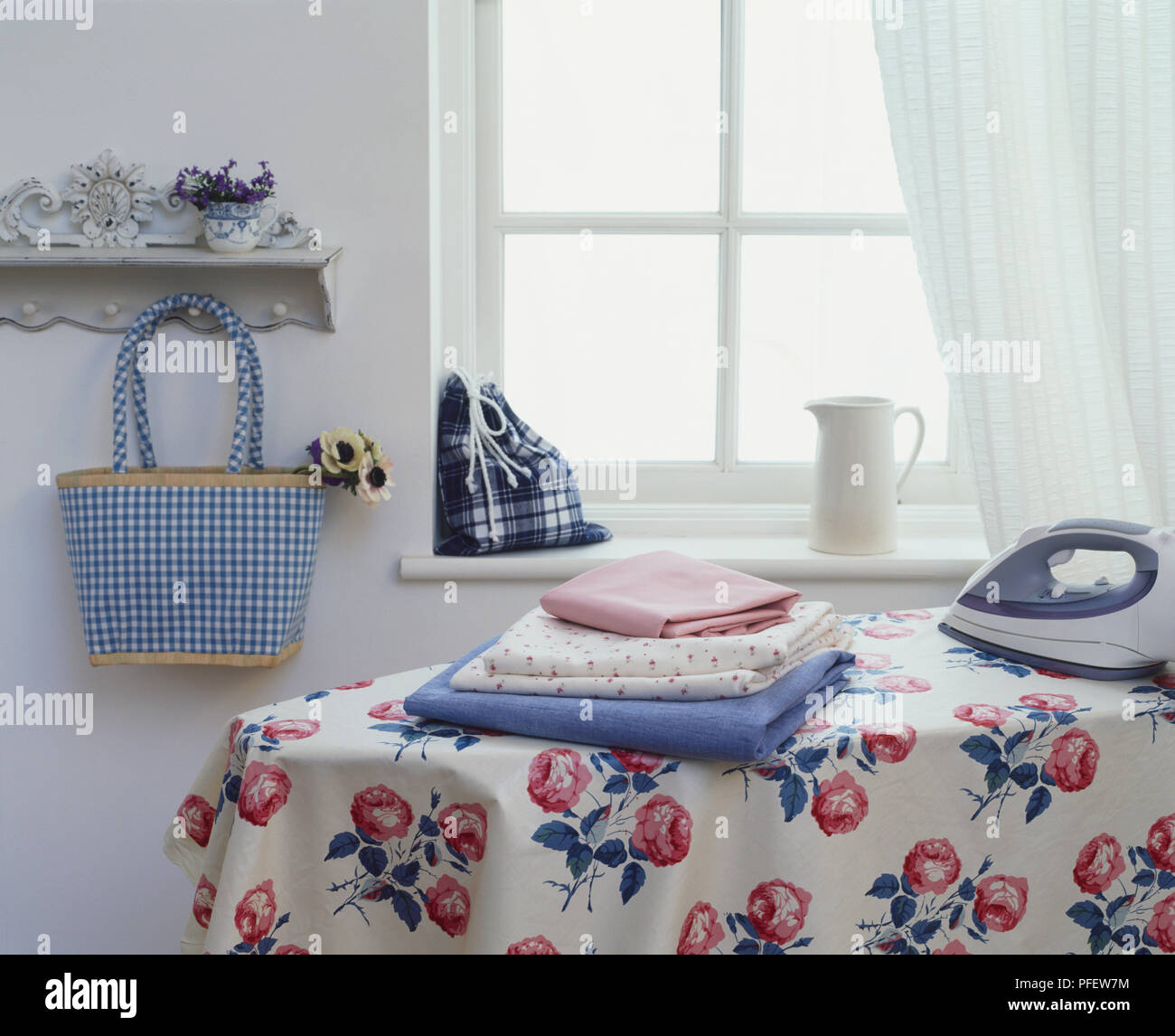 Rose patterned material draped over ironing board, folded fabrics and iron placed on top, chequered and tartan bags displayed in background, side view Stock Photo