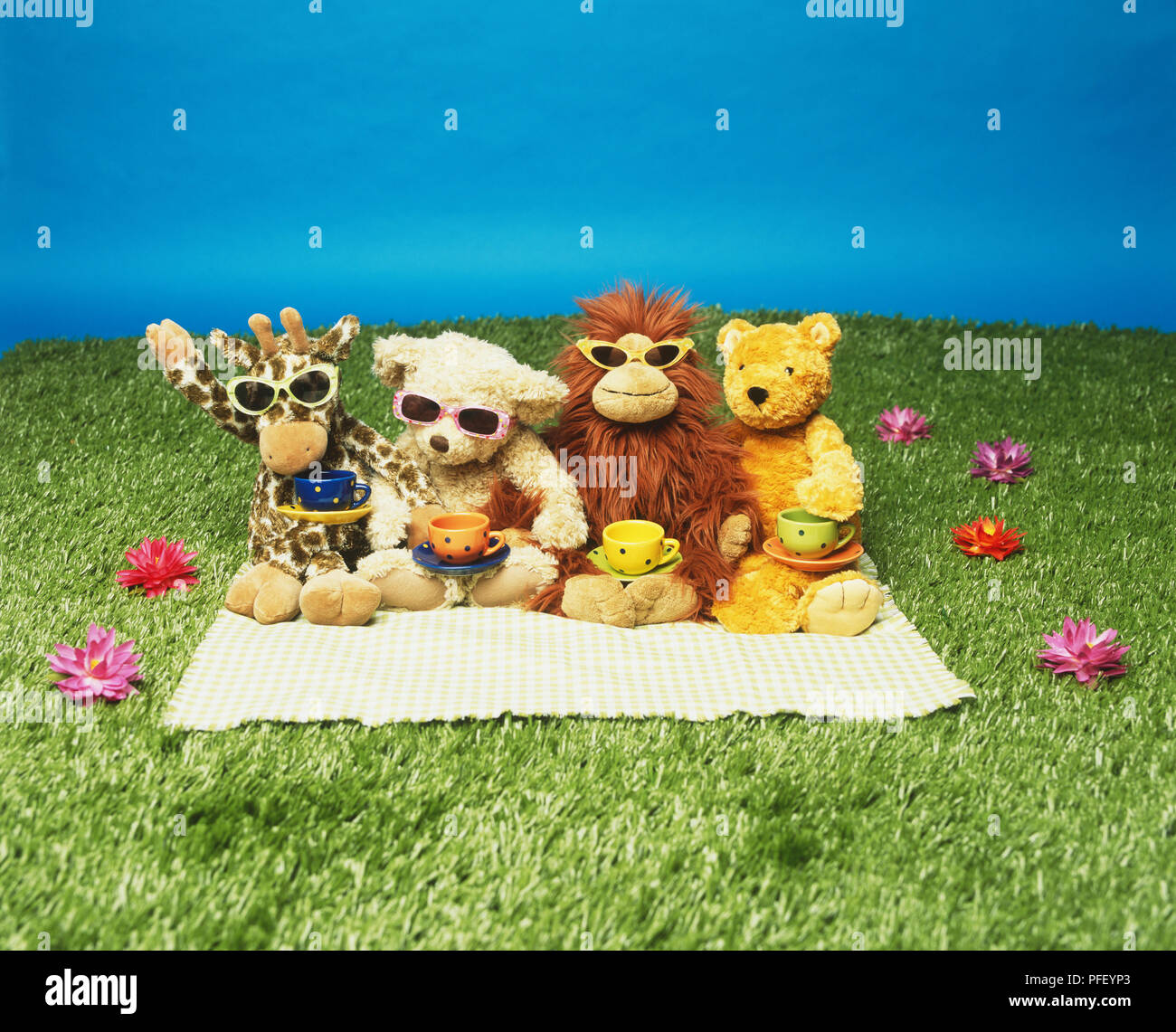 Toy giraffe, two bears and lion with sunglasses on, tea cups on their laps, sitting on a blanket in the grass Stock Photo
