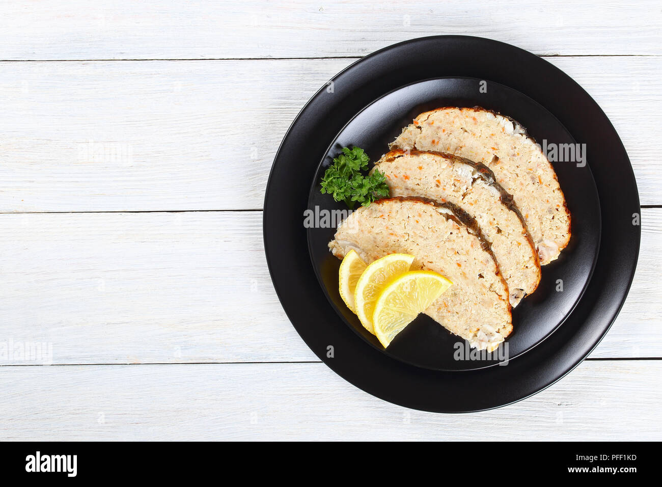 delicious baked in oven Gefilte minced carp fillets cut in slices with fresh parsley, lemon on black plate,  on white wooden table, view from above, c Stock Photo
