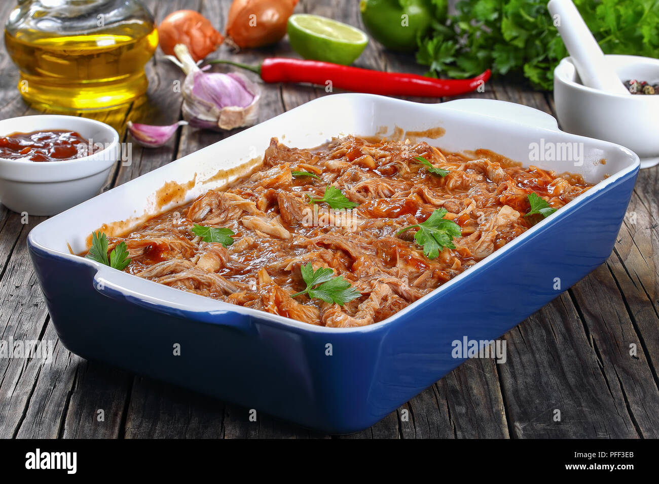 Shredded chicken meat tossed in barbecue sauce, loaded with smoky chipotle flavours in baking dish, on old wooden table with ingredients on background Stock Photo