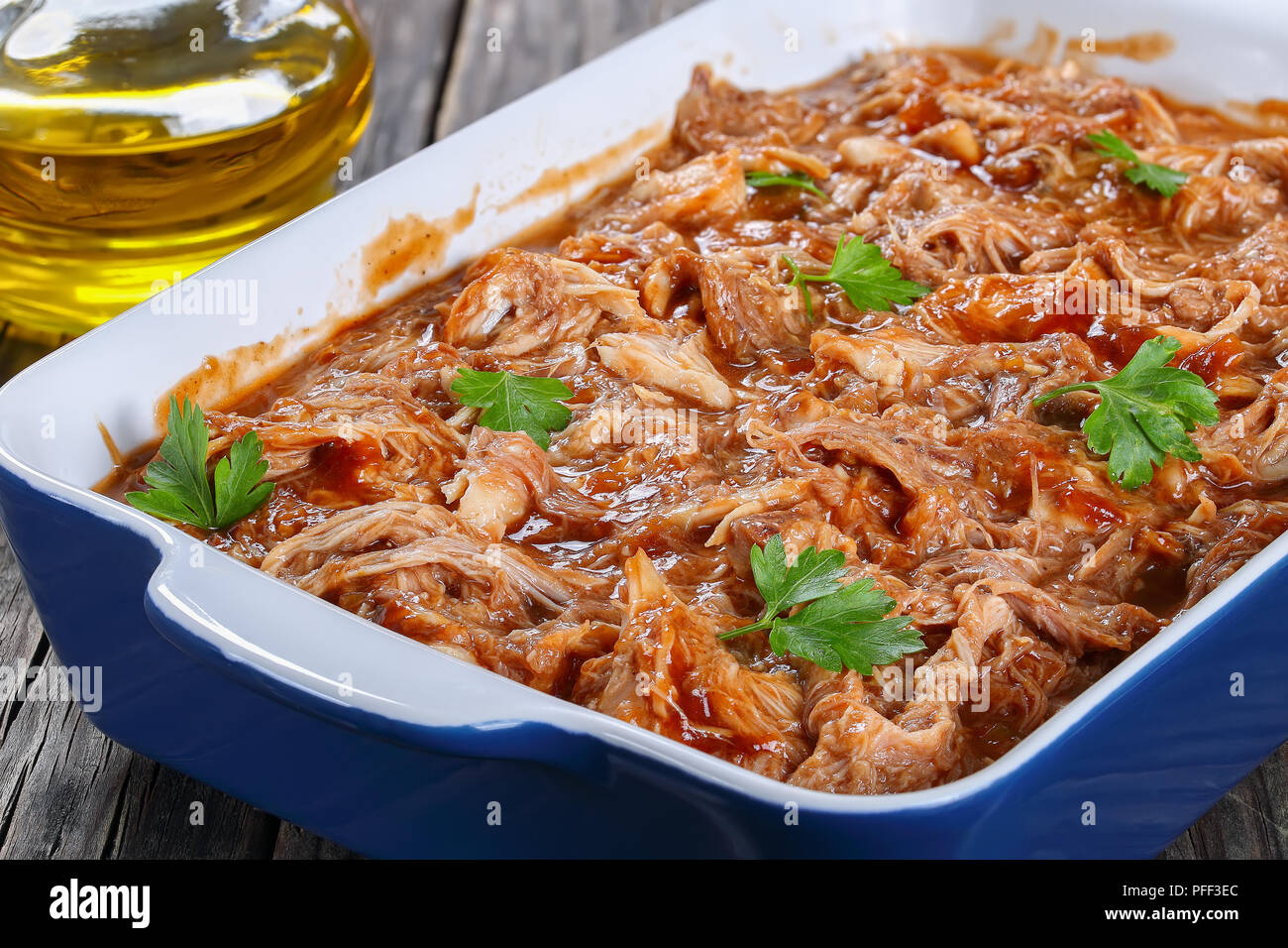 delicious Shredded chicken meat tossed in barbecue sauce, loaded with smoky chipotle flavours in baking dish, on old wooden table with olive oil on ba Stock Photo
