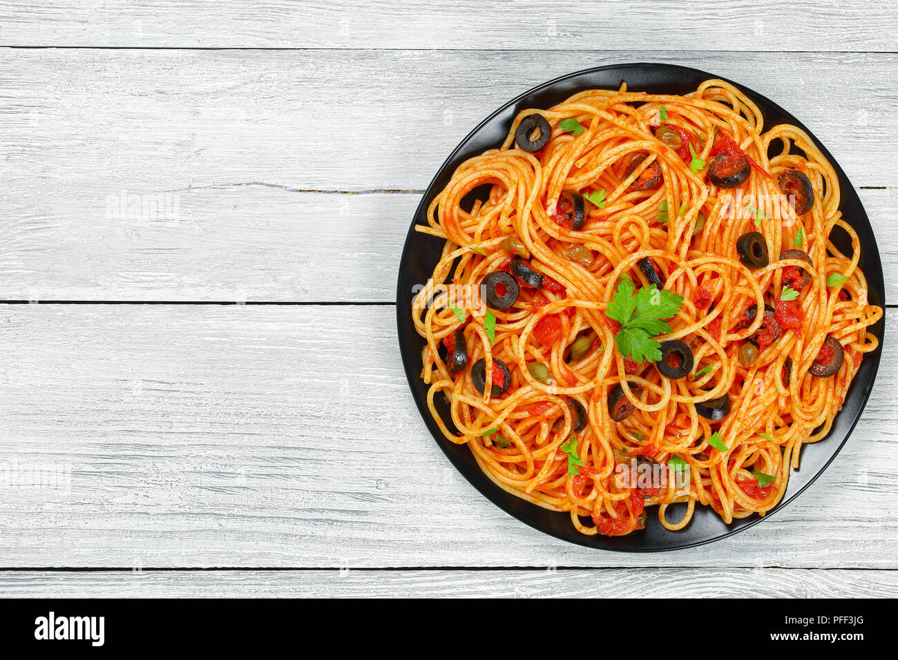 Delicious Spaghetti alla puttanesca with capers. olives, tomato sauce sprinkled with parsley on black plate on wooden table, authentic basic recipe, v Stock Photo