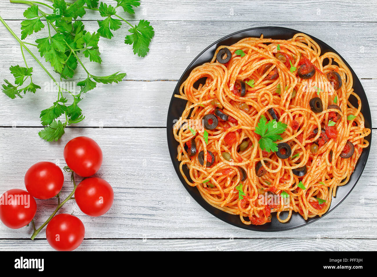 Delicious Spaghetti alla puttanesca with capers. olives, anchovies, tomato sauce sprinkled with parsley on black plate on wooden table, authentic basi Stock Photo