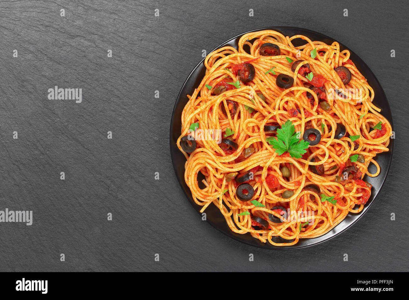 Delicious Spaghetti alla puttanesca with capers. olives, anchovies, tomato sauce sprinkled with parsley on black plate on stone tray, authentic basic  Stock Photo