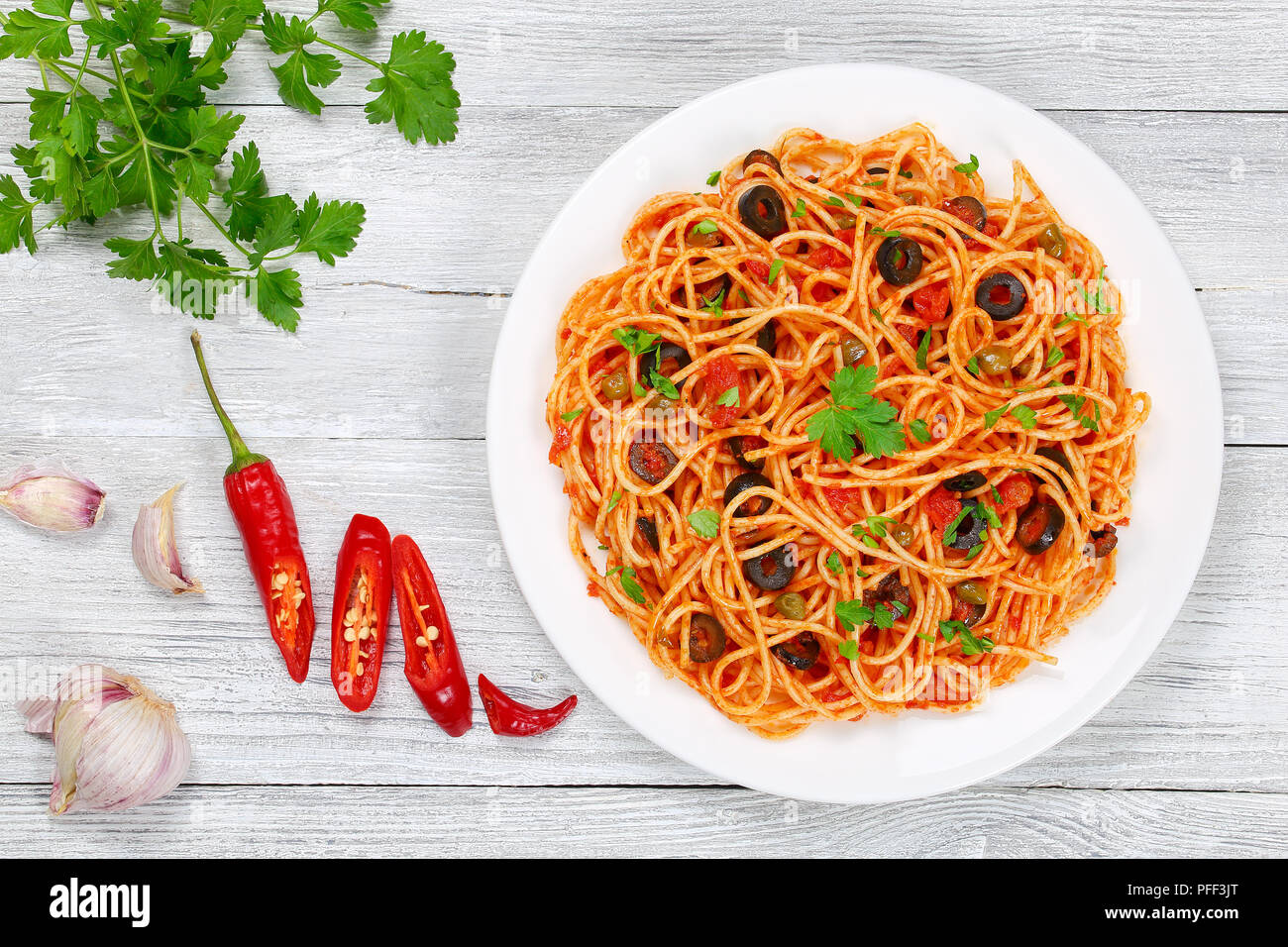 Delicious Spaghetti alla puttanesca with capers, olives, anchovies, tomato sauce sprinkled with parsley on white plate on wooden table with ingredient Stock Photo