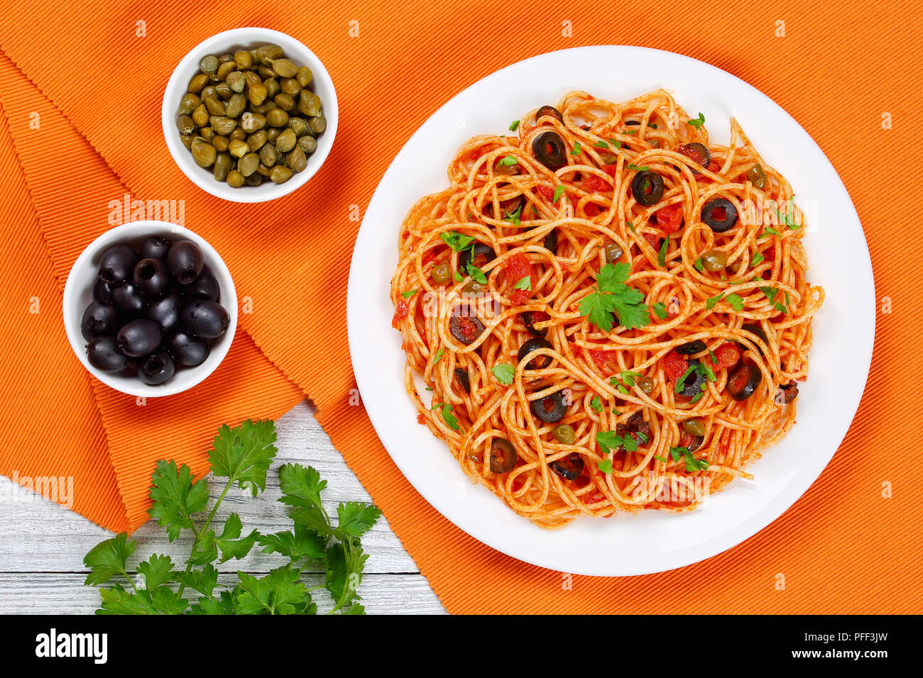 Delicious Spaghetti alla puttanesca with capers, olives, anchovies, tomato sauce sprinkled with parsley on white plate on table mat with olive and cap Stock Photo