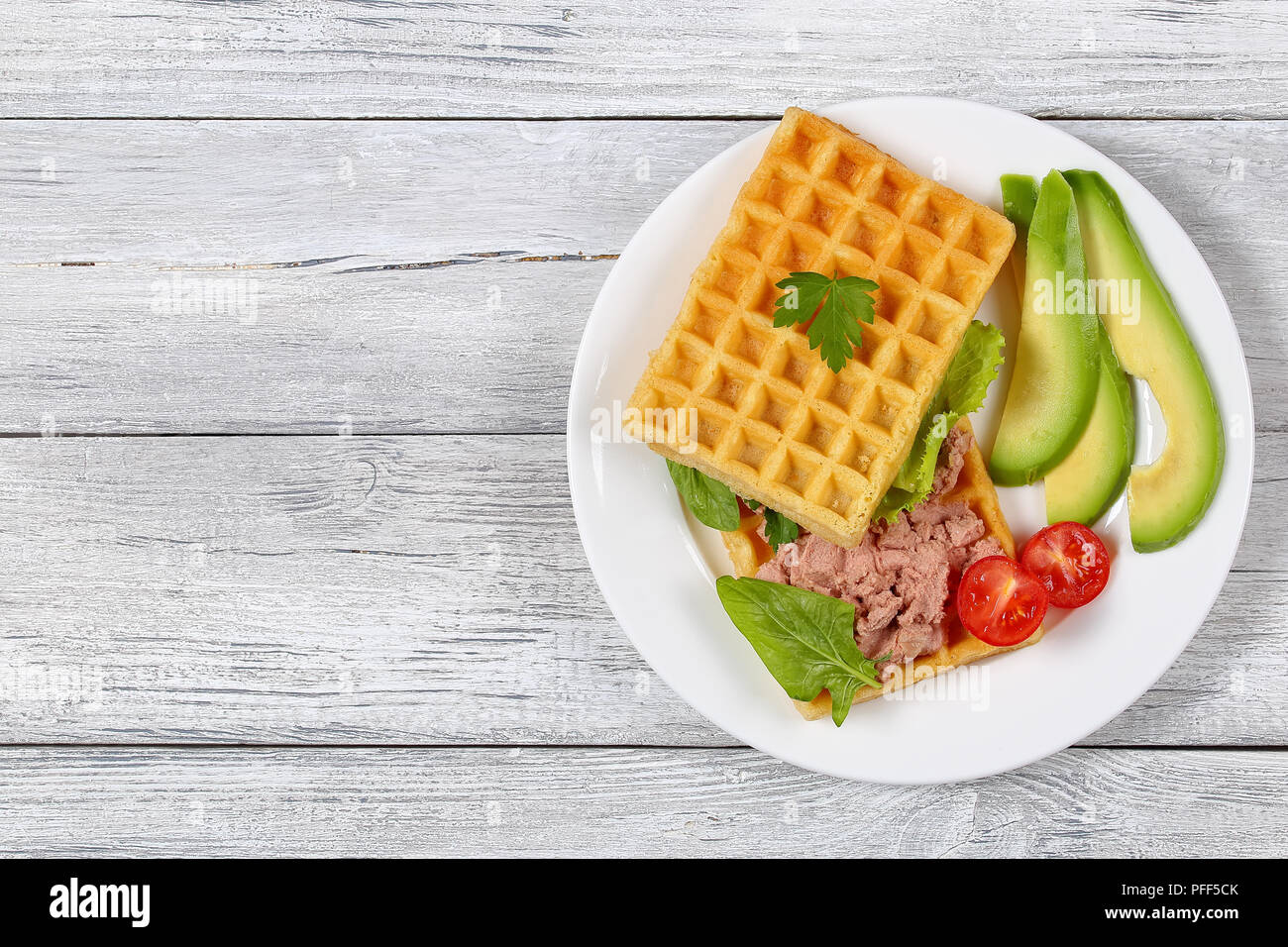 delicious salted belgian waffles sandwich with avocado slices, turkey liver pate, lettuce, spinach and parsley on white plate with cherry tomatoes, vi Stock Photo