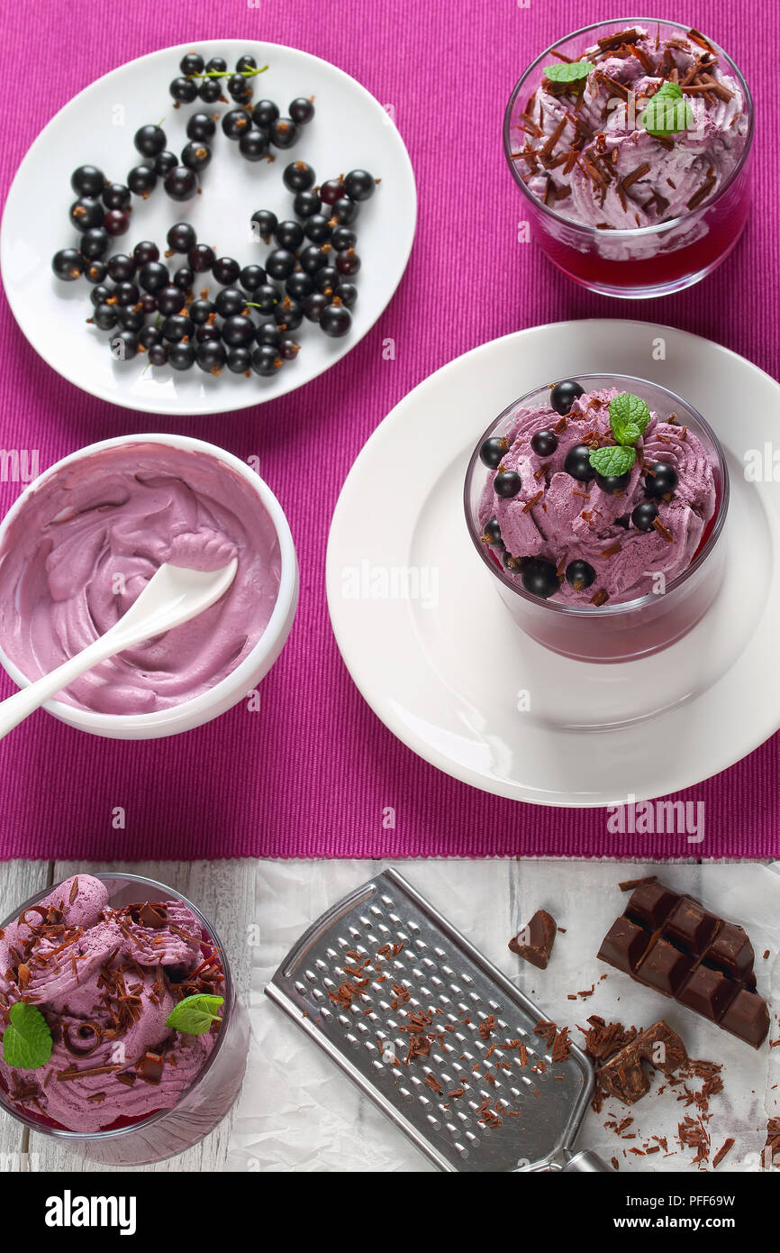 homemade ice cream dessert of blackcurrant, ricotta, yogurt mousse decorated with mint leaves in glass cups on plate.chocolate bar and grater on paper Stock Photo