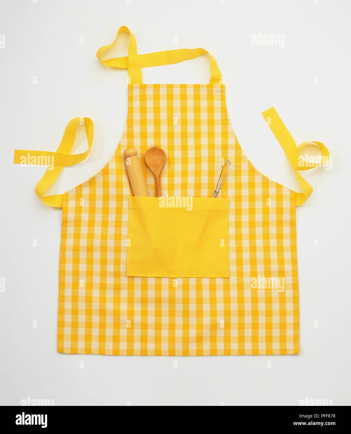 Chequered yellow apron with wooden spoon, rolling pin and whisk in the front pocket, front view. Stock Photo