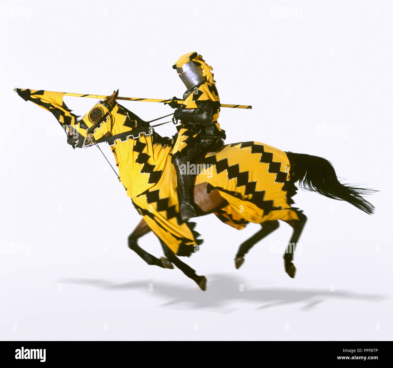 Knight charging ahead on horseback with pointed spear, horse draped in yellow and black fabric matching knight's uniform, side view. Stock Photo