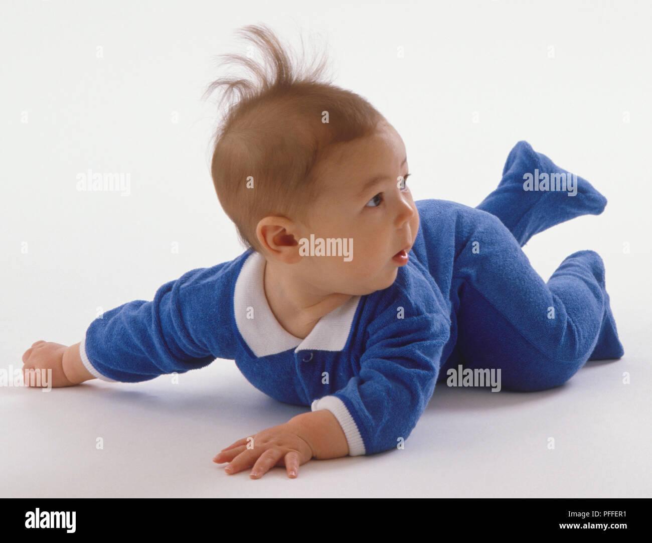 Baby wearing blue and white jumpsuit, hair standing on end, crawling on his belly. Stock Photo