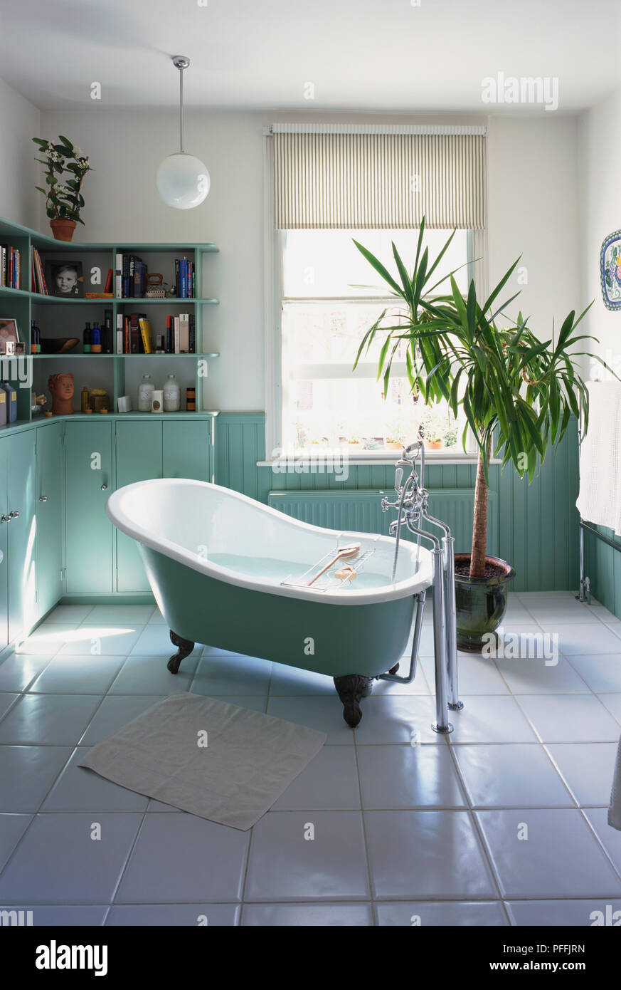 Large bright bathroom; freestanding white and green bath with iron feet, wooden wall panelling painted green, cupboards and tall shelves, tiled floor, potted Yucca, open window. Stock Photo
