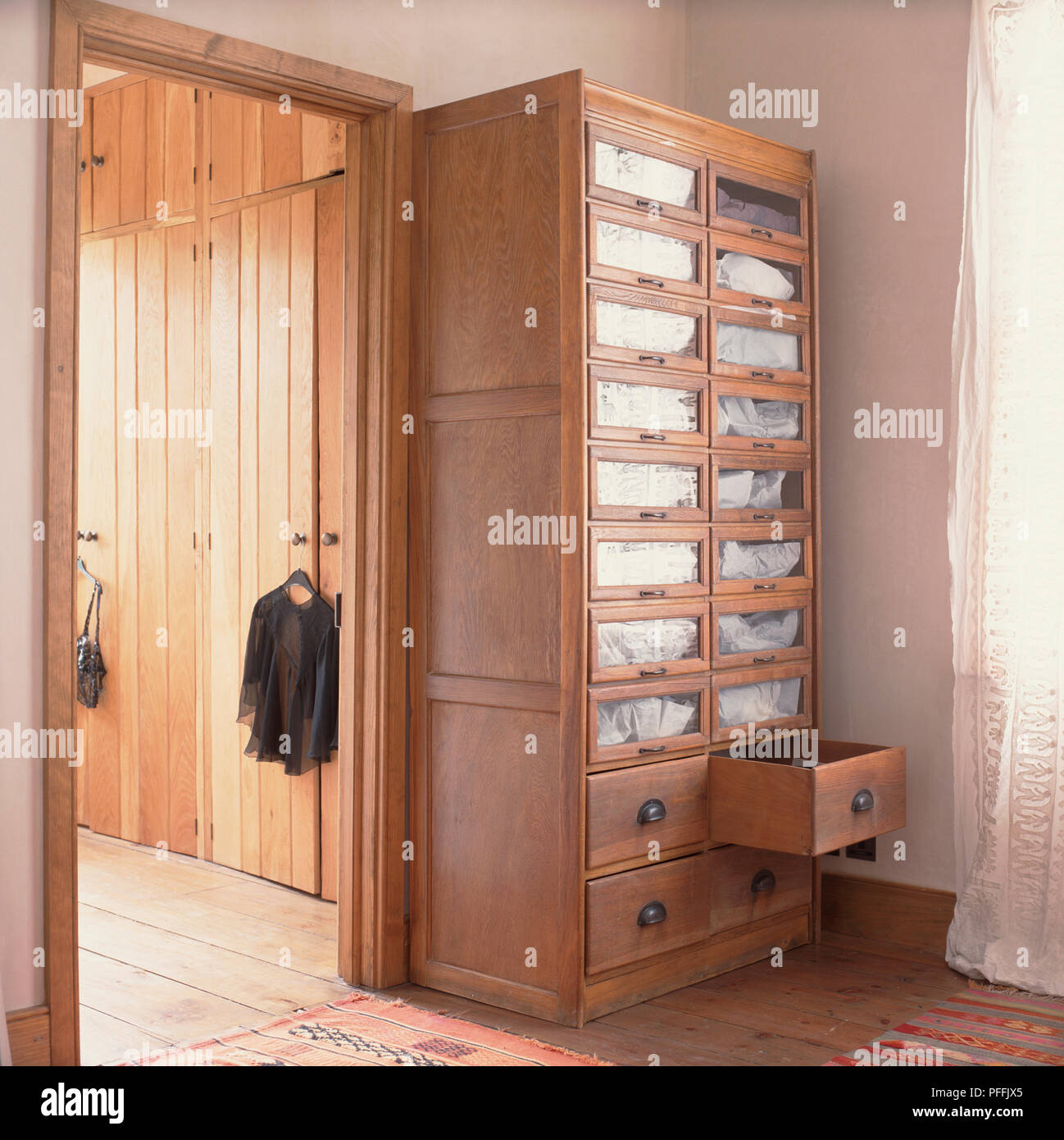 Cupboard-like wooden shelving unit, large drawers at bottom, small glass-fronted drawers reaching to top, white material behind glass windows, wooden-floored room with large wooden wardrobes through doorway. Stock Photo
