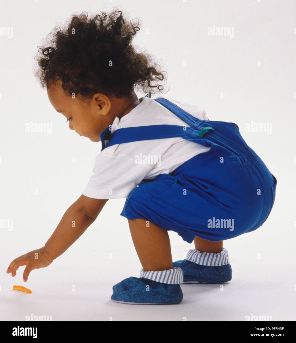 Toddler wearing blue dungarees, squatting and reaching down to pick up a sweet from the floor. Stock Photo
