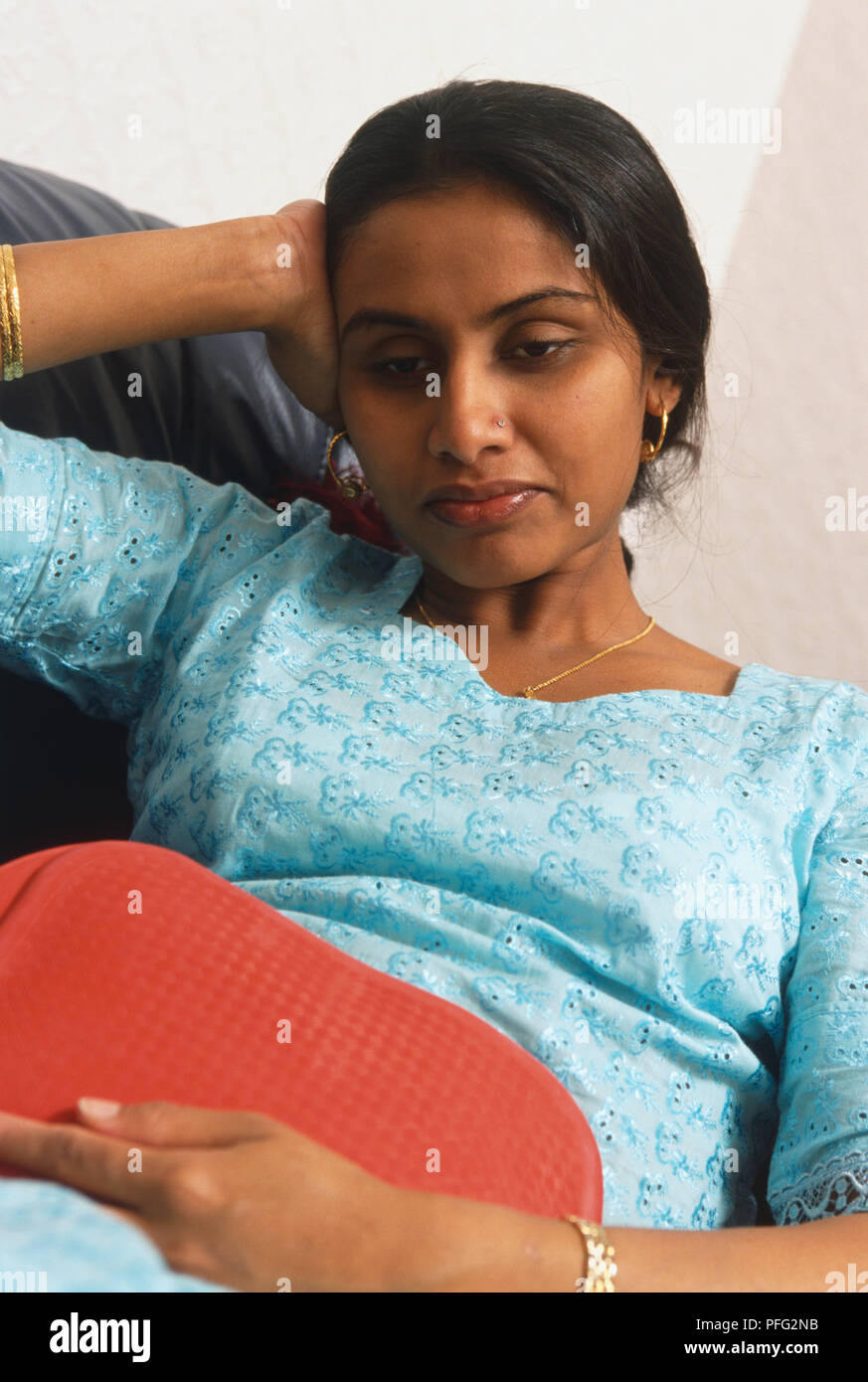 Sitting woman leaning her head on one hand and holding a hot water bottle to her belly with the other. Stock Photo