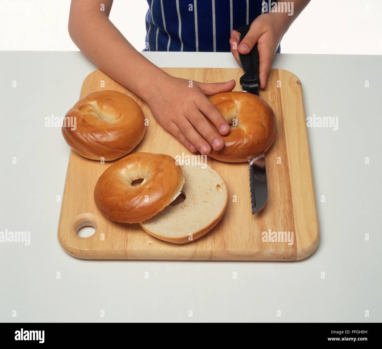 Boy using kitchen knife to cut bagel in half on wooden Choppingboard Stock Photo