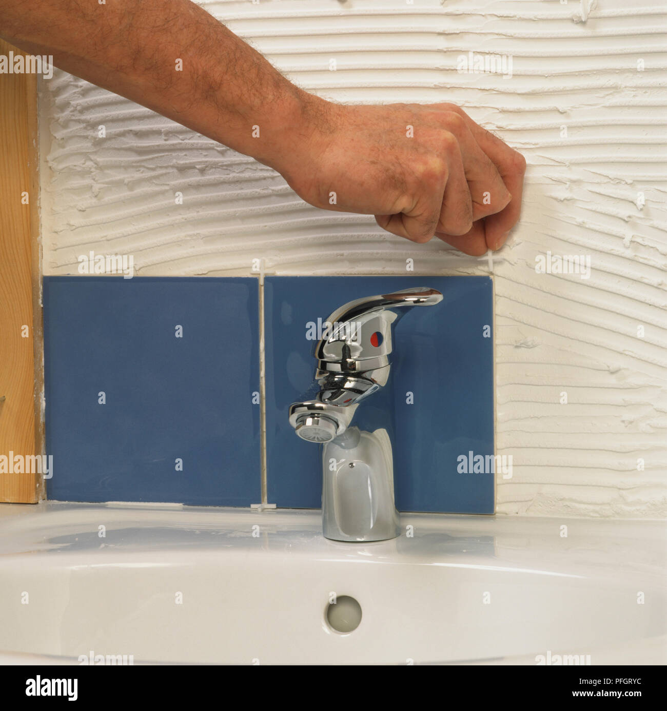Hand placing a tile spacer on a bathroom wall covered in adhesive, two blue tiles already laid ...