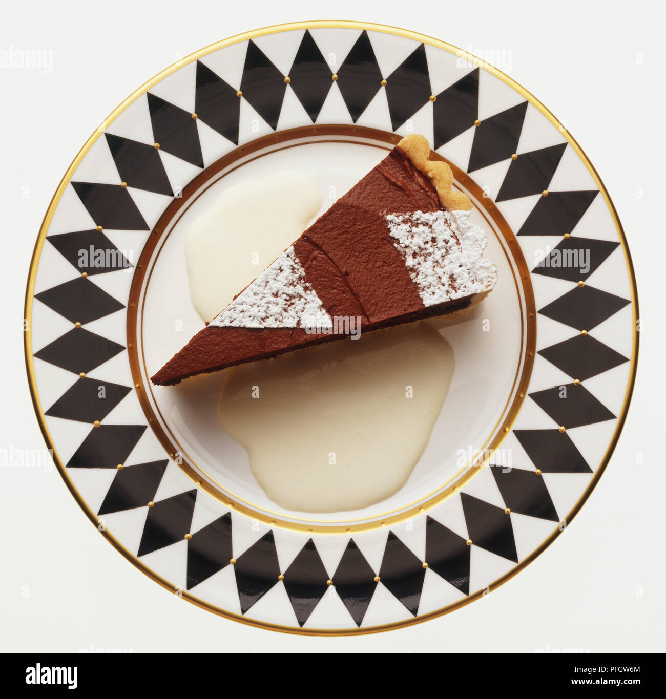 A piece of chocolate tart sprinkled with icing sugar, served with cream on a decorative black and white patterned plate, view from above Stock Photo