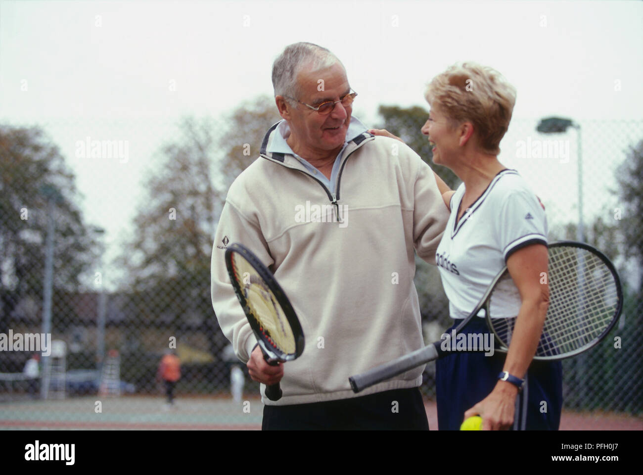 Mature man holding tennis racket, mature woman with racket under arm and tennis ball in hand, each smiling and with arm around other, on tennis court. Stock Photo