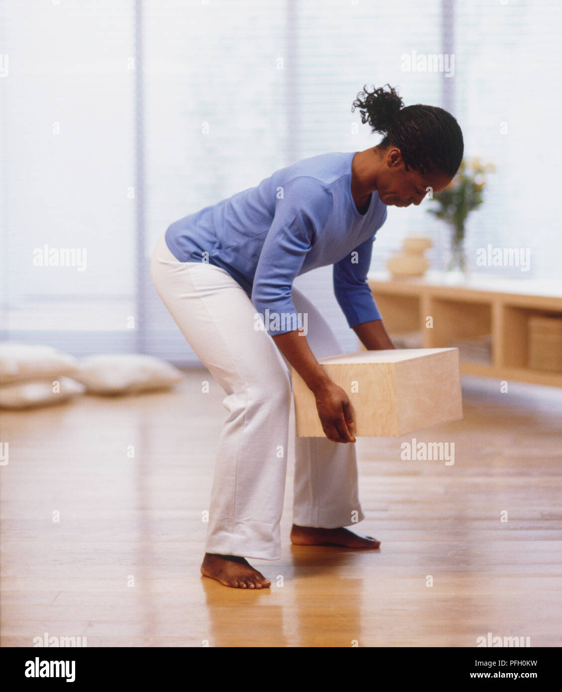Woman in blue shirt and white trousers lifting box off floor, side view