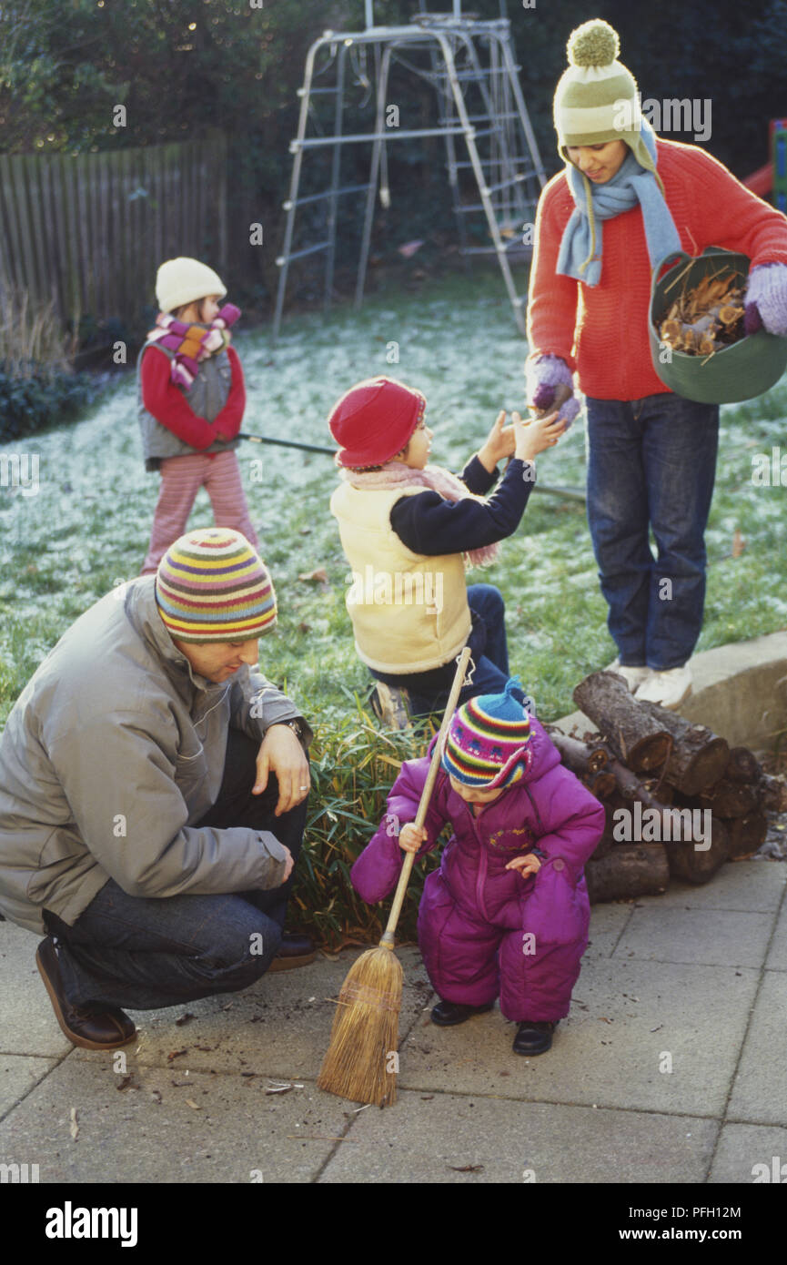 Family in garden, man kneeling next to boy toddler sweeping with broom, woman handing stick to girl from bag of leaves, girl in background raking Stock Photo