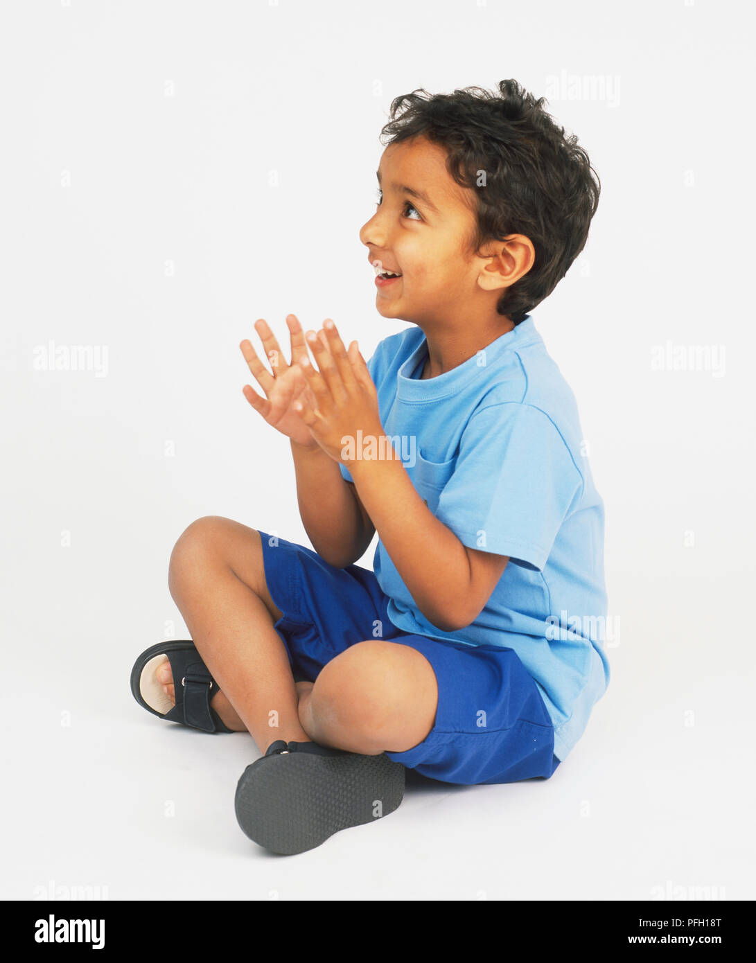 Boy sitting cross legged on floor, looking up and clapping hands Stock Photo