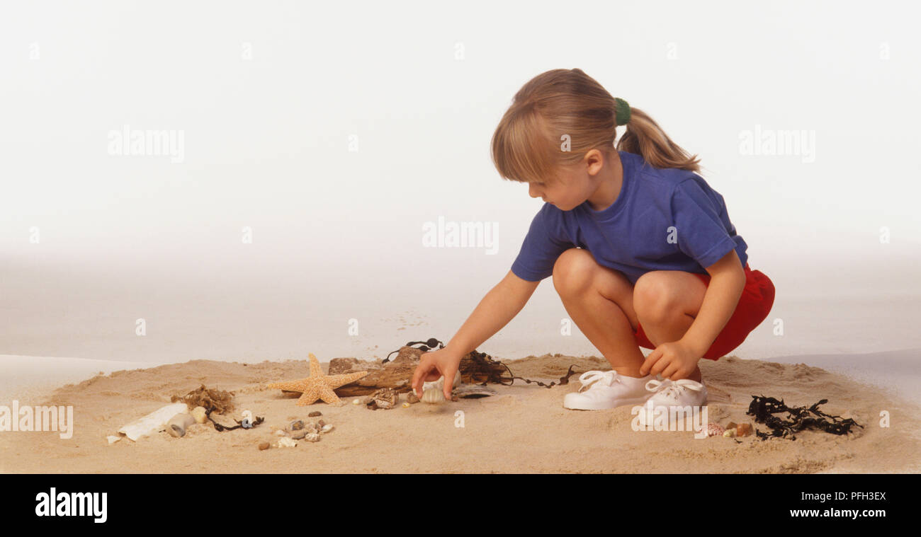 Crouching Blonde Girl Arranging Seashells On Sand Side View Stock