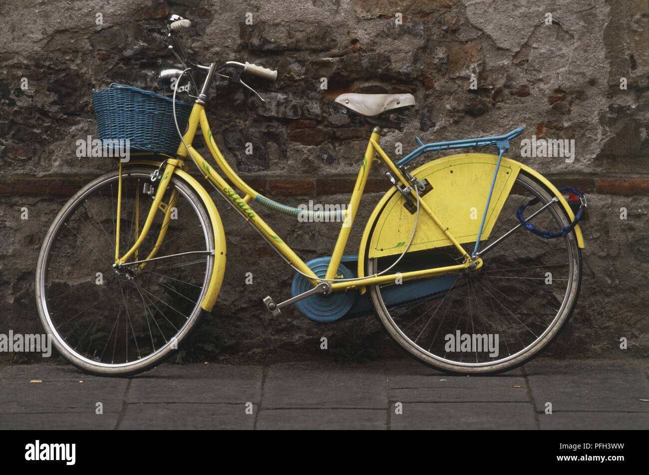 Yellow women's bicycle with a wicker basket mounted on front wheel, leaning against a weathered old wall Stock Photo