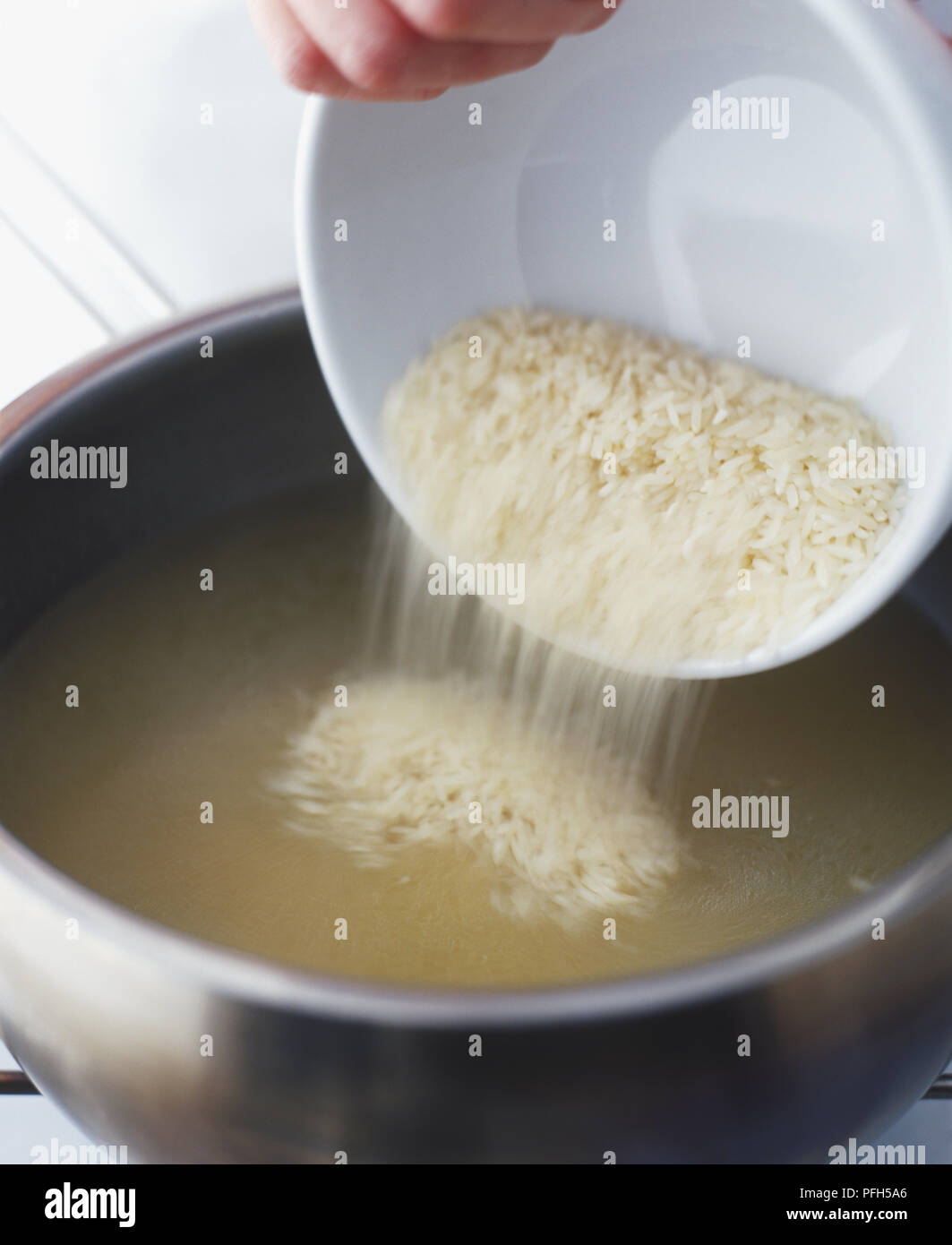 Pouring rice from a bowl into a pan of chicken stock Stock Photo