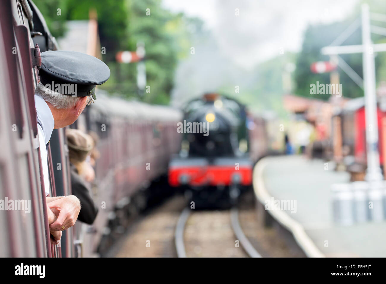 Rear view of ticket inspector (taken on board) leaning out of vintage carriage as train approaches station. Second steam engine is  approaching. Stock Photo