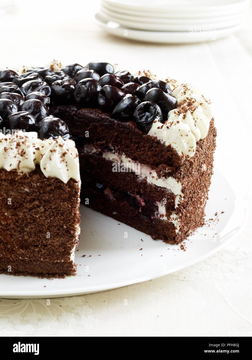 Black Forest Gateaux, decorated with chocolate, cherries and cream, with a slice cut away, stack of plates nearby, close-up Stock Photo
