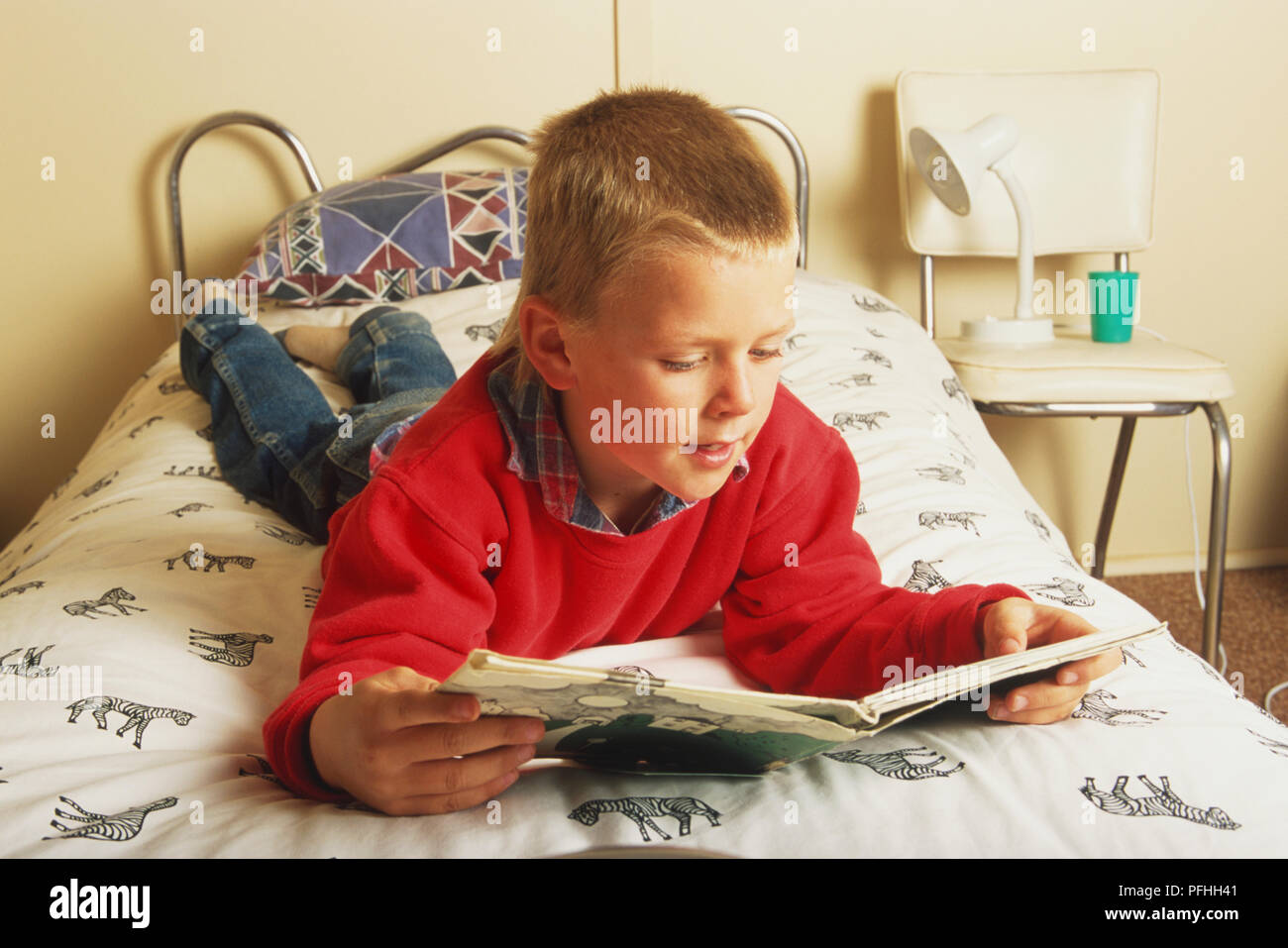 Blonde boy lying on his front on a bed reading a book, front view. Stock Photo