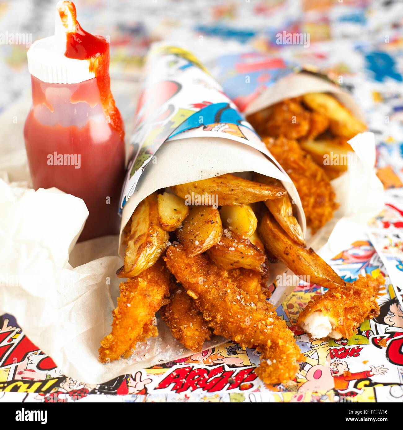 Deep-fried pieces of fish and chips in paper cone, next to bottle of tomato ketchup Stock Photo
