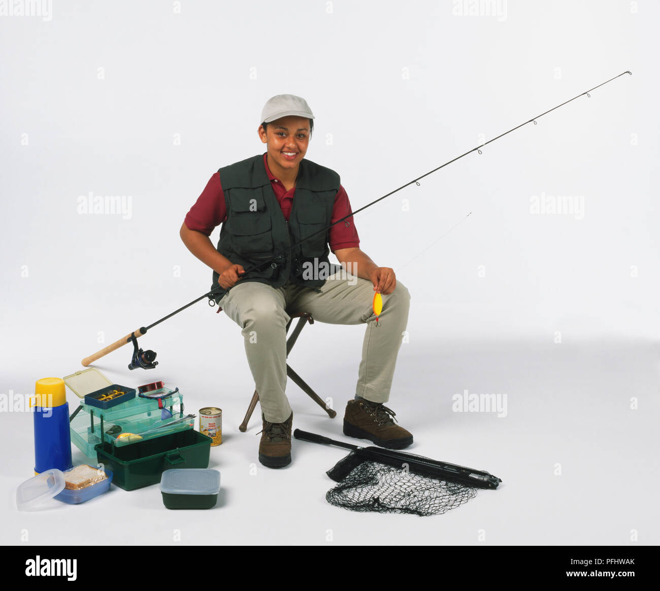 https://c8.alamy.com/comp/PFHWAK/smiling-teenage-girl-in-waistcoat-and-peaked-cap-sitting-on-stool-holding-fishing-rod-in-one-hand-and-plug-in-the-other-surrounded-by-fishing-gear-scattered-on-floor-front-view-PFHWAK.jpg