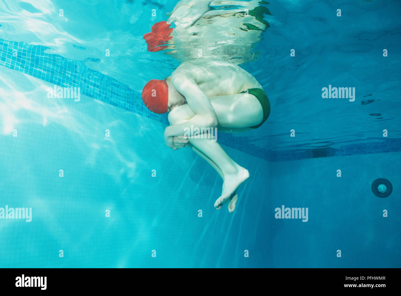 Teenage boy in green swimming trunks and red cap curling forward in foetal position under water, low angle view Stock Photo