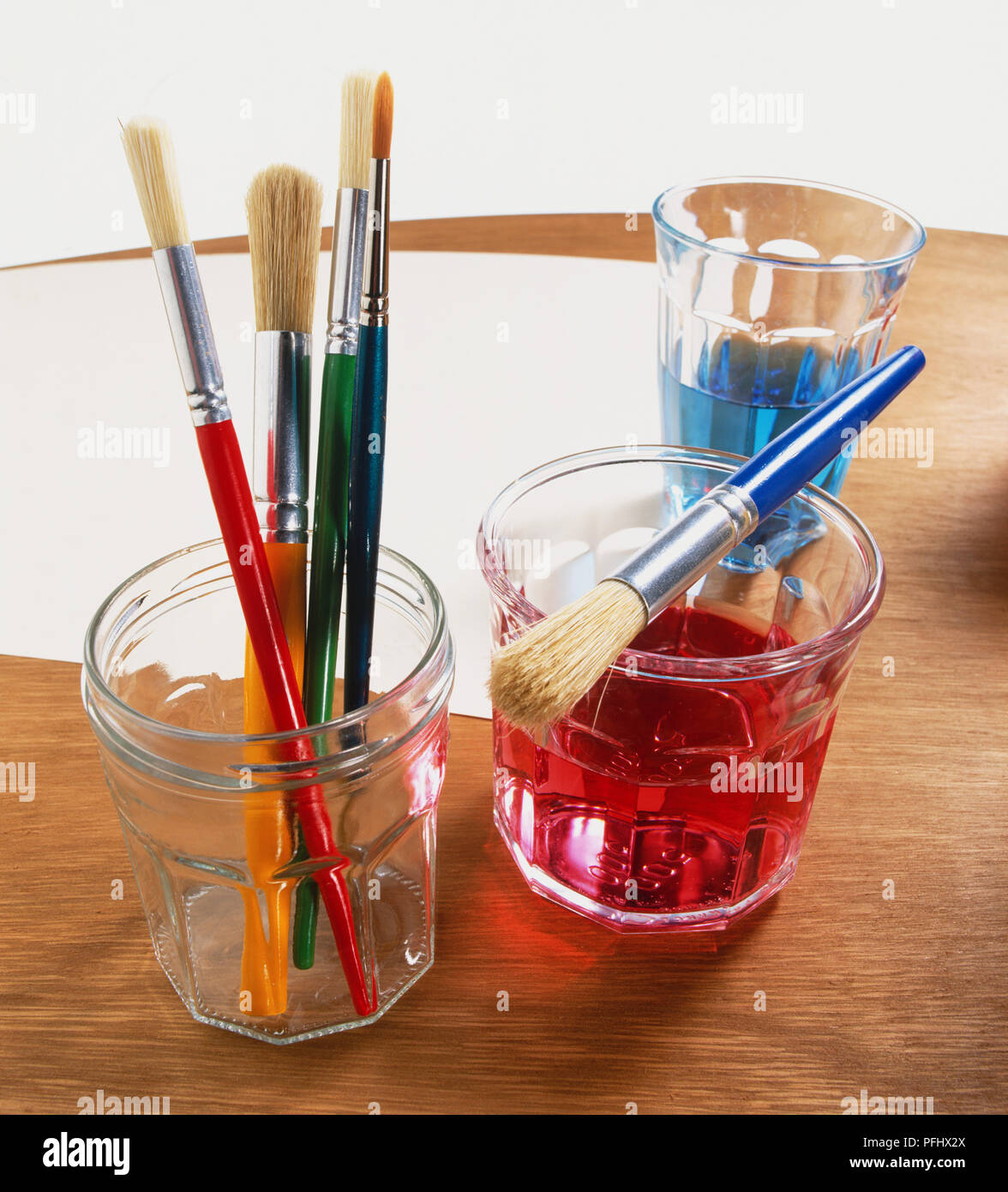 Glass tumbler with red coloured water and paint brush resting on rim, tumbler with blue water, assorted paint brushes in a jar, sheet of paper on table, front view. Stock Photo