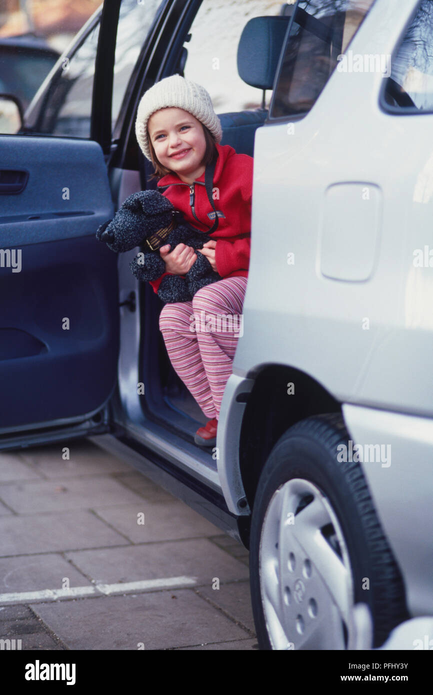 Smiling girl wearing a wooly hat looking out from the back of a car with its door open. Stock Photo