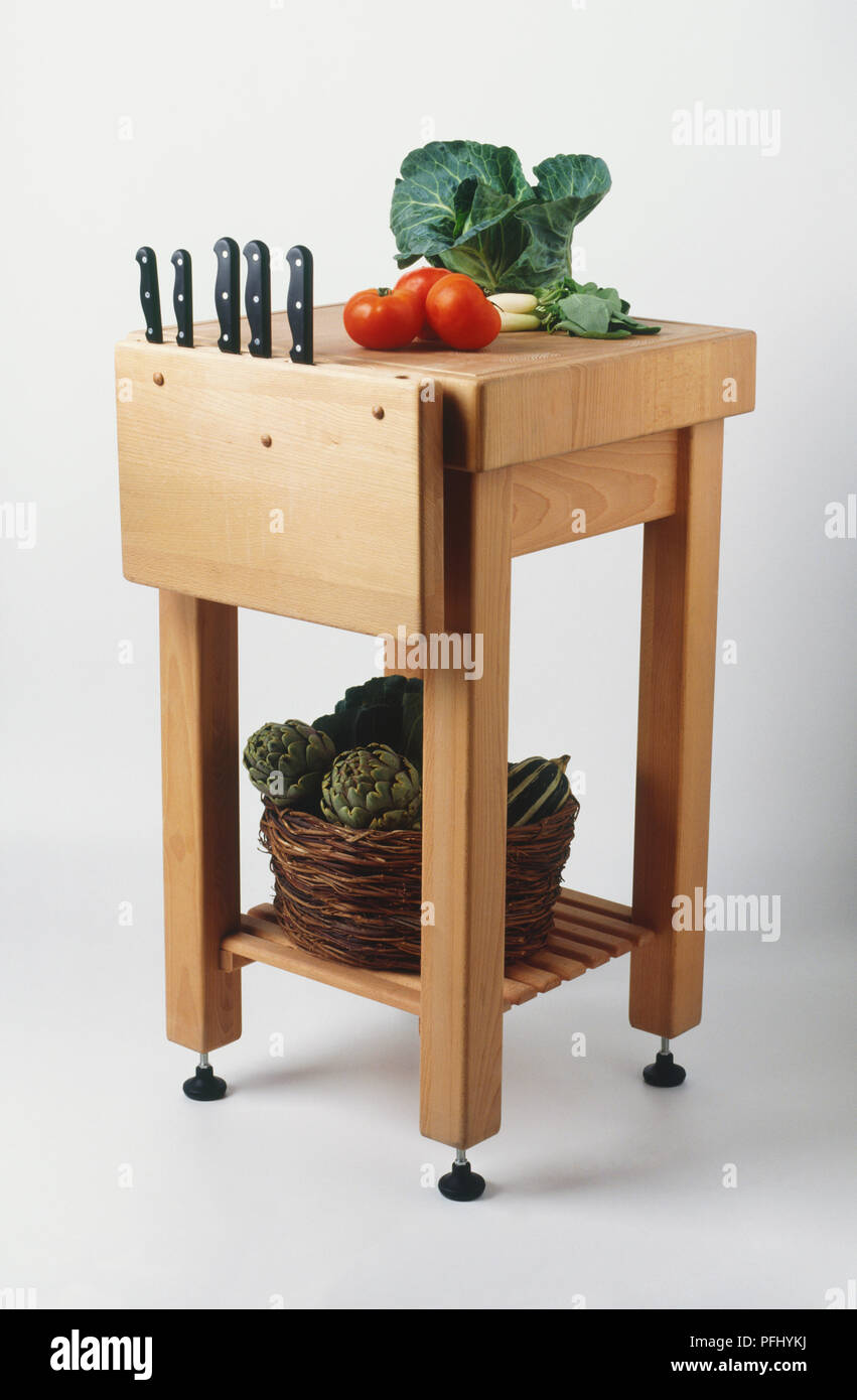 Butcher's block, shelf at bottom holding basket of artichokes, cabbage and tomatoes on top, set of knives inserted into side block. Stock Photo