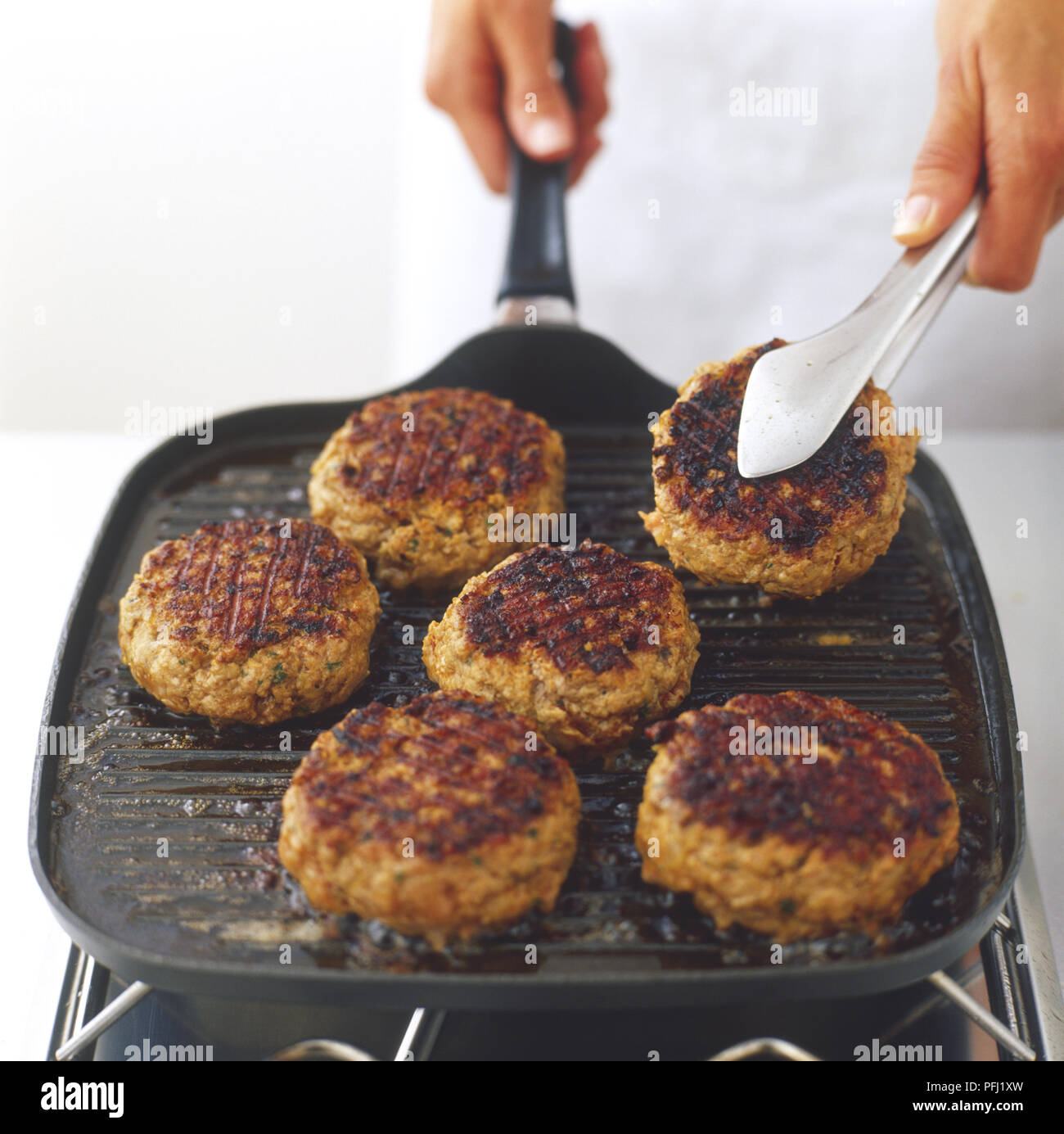 Six burgers grilling in a grill pan, one being turned over with tongs