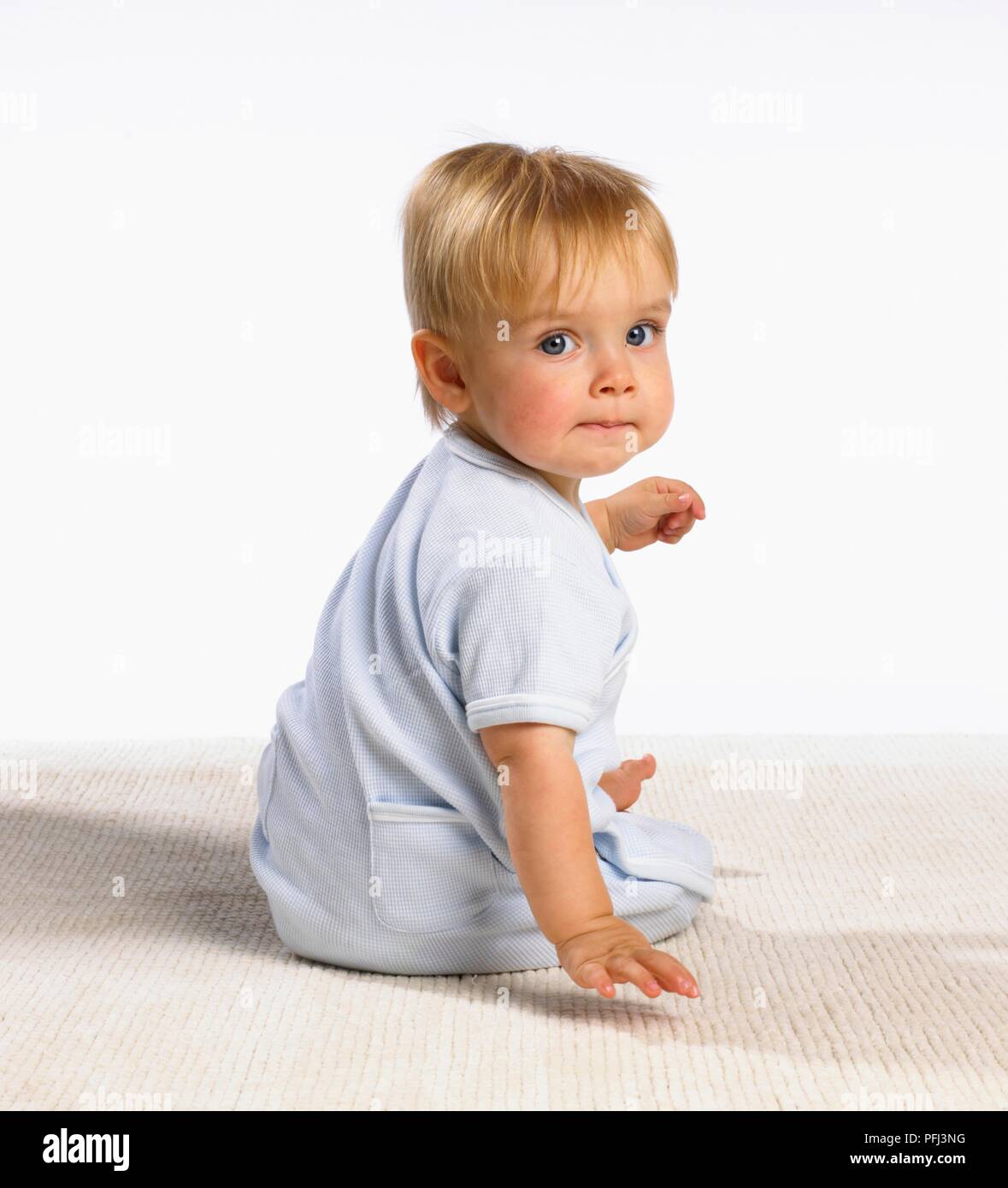Blonde baby boy in blue pyjamas sitting on floor, looking over shoulder