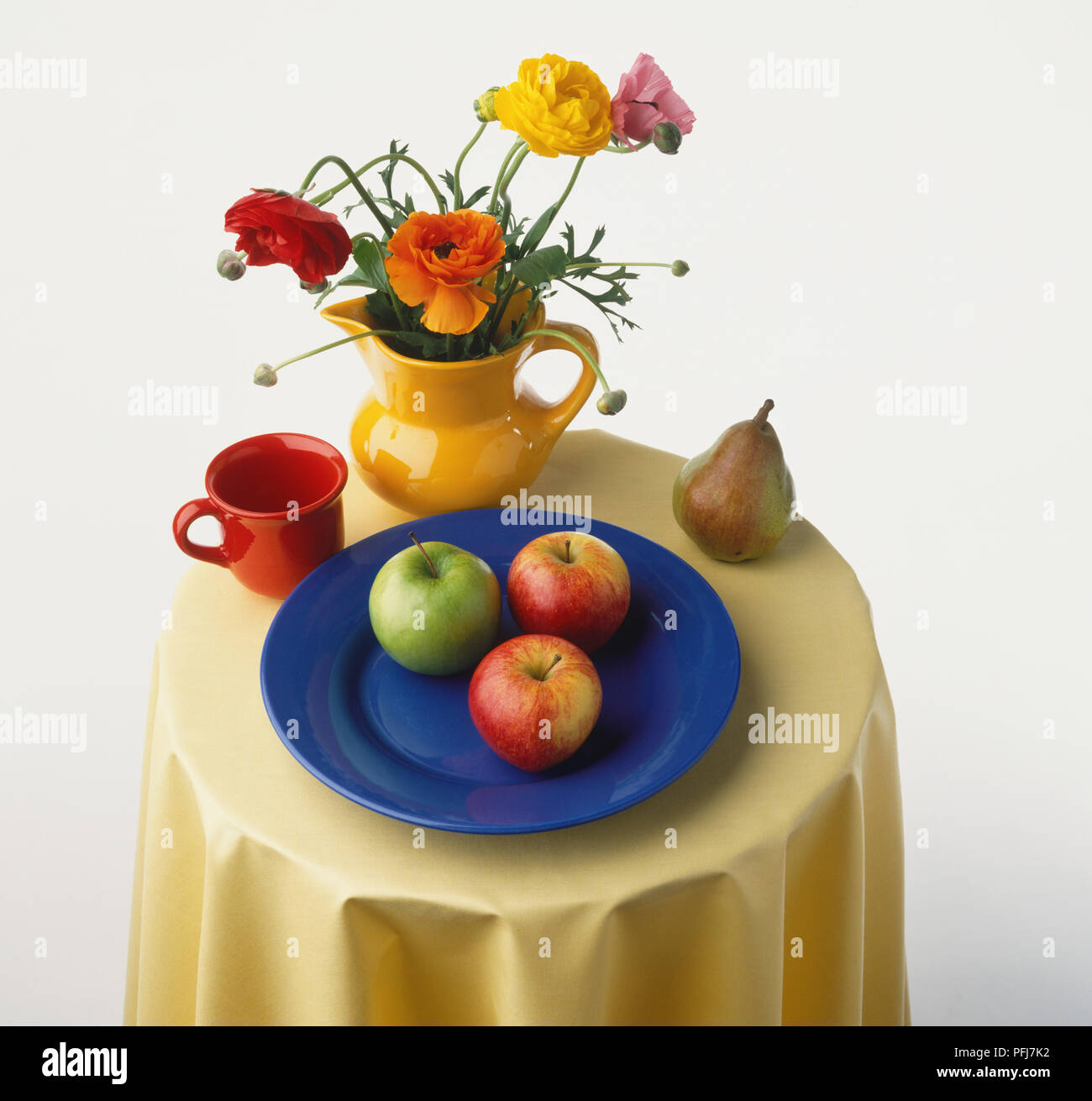 Small round table laid with red mug, colourful flowers in yellow jug, three apples on blue plate, pear,  elevated view. Stock Photo