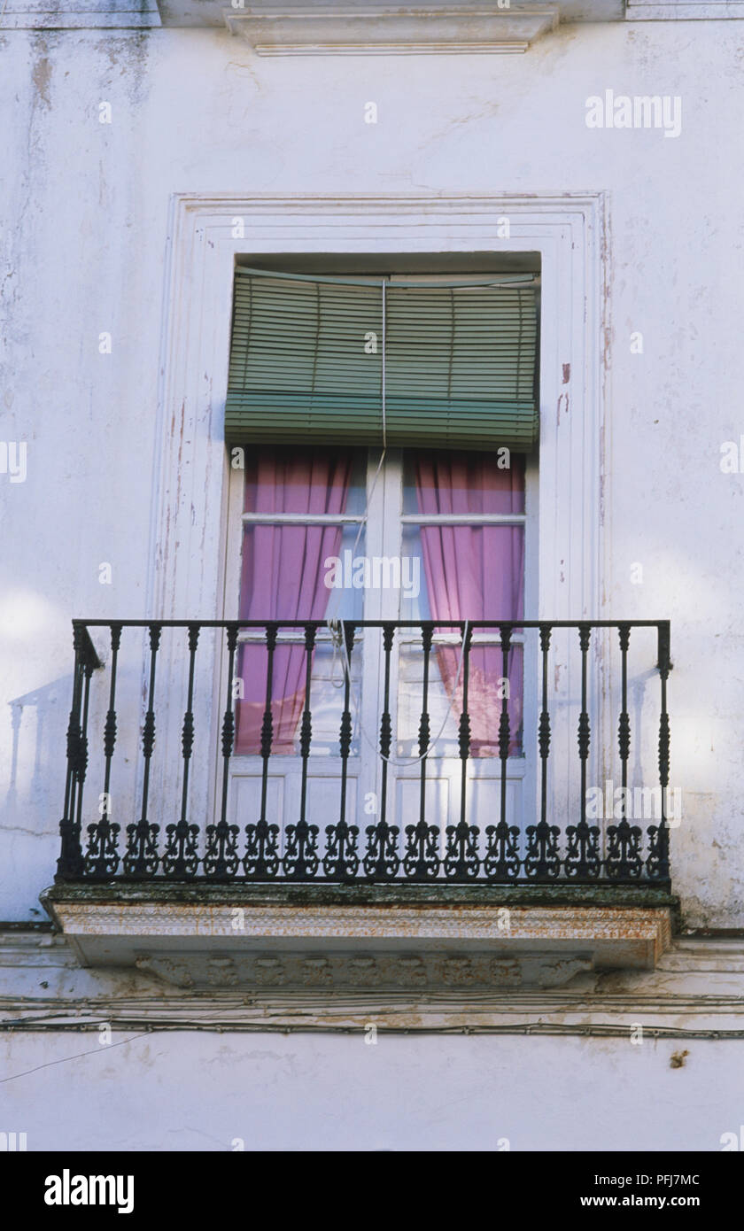 Outside view of narrow balcony with iron railings, door with white window frames, purple curtains tied to side, half-closed green blinds, white wall with worn off white paint. Stock Photo