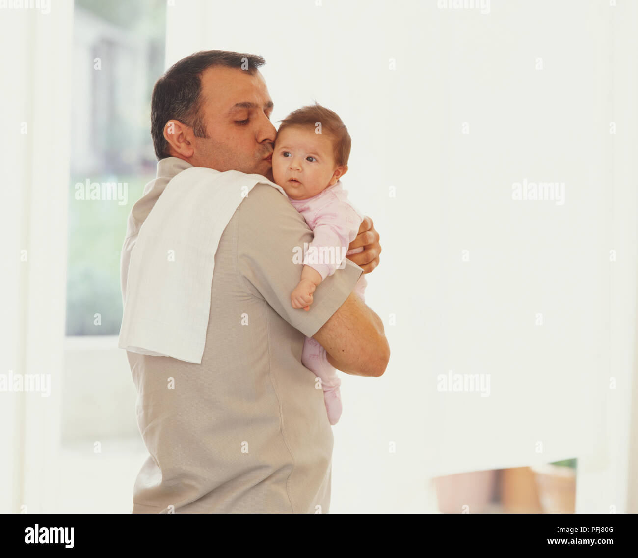 Baby looking over the shoulder of man who is holding her, and planting a kiss on her cheek Stock Photo