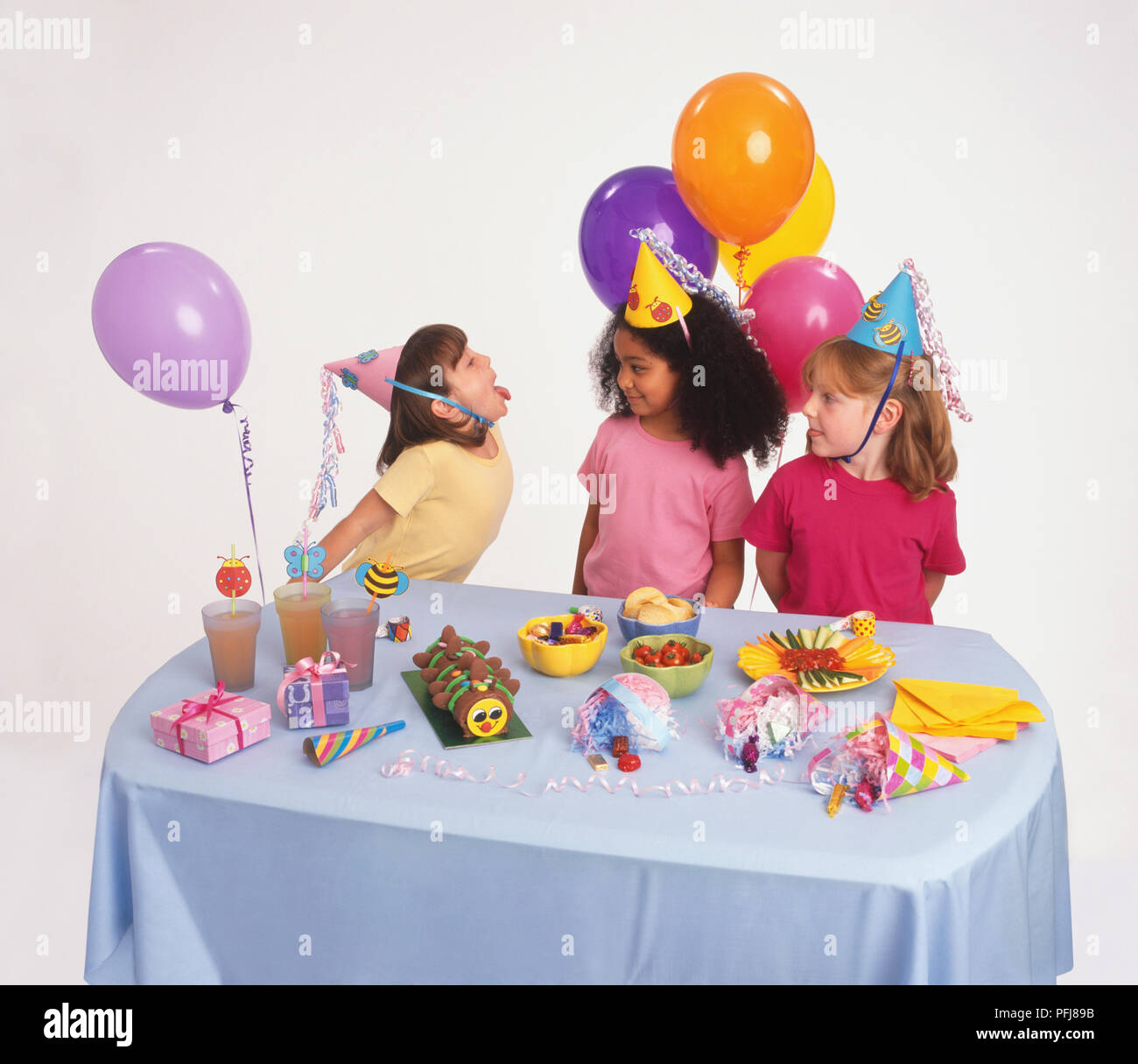 Girl in party hat sticking tongue out at two other girls, all standing behind table decorated for birthday party. Stock Photo