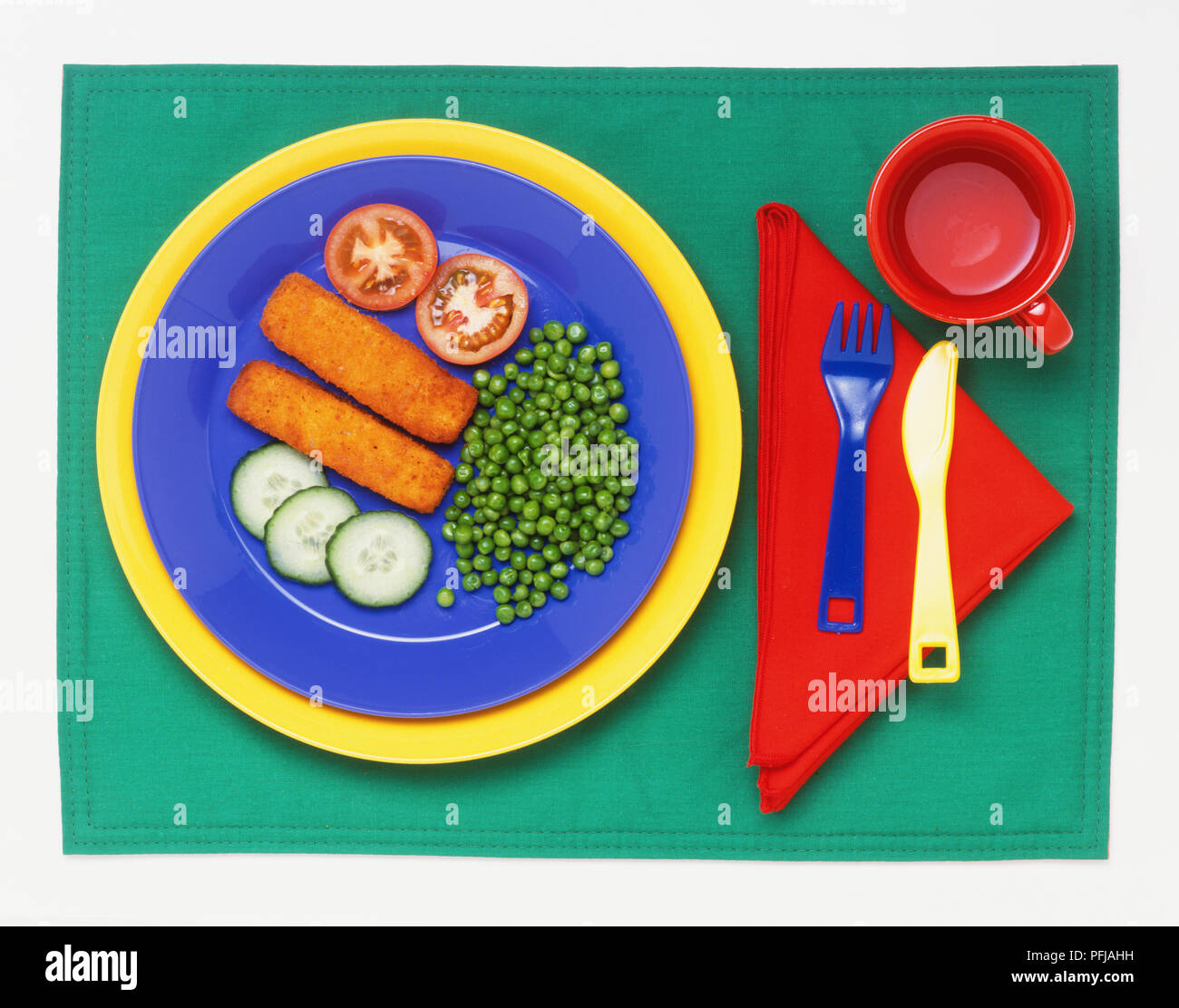 Fish fingers, peas, tomato and cucumber slices on a plate, plastic fork and knife on folded red napkin, and red mug, view from above Stock Photo