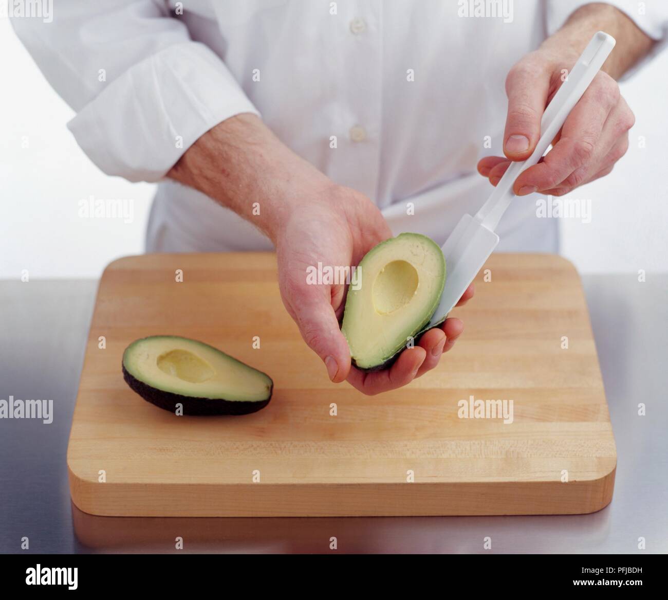 Holding avocado half in one hand and using rubber spatula to scoop out flesh, second avocado half on chopping board Stock Photo